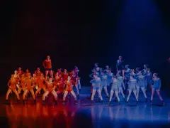 Grupa tancerzy na scenie, podzielona na dwie grupy: jedna w pomarańczowych strojach, druga w niebieskich. Nauka tańca nowoczesnego w pełnym blasku.
