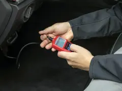 Ręce trzymające czerwony interfejs diagnostyczny OBDII/EOBD, podłączony do samochodu.
