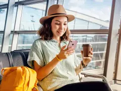 Kobieta w kapeluszu siedzi na lotnisku z kawą i telefonem, martwiąc się o brak zasięgu za granicą Play.