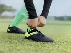 Dłonie wiążą sznurówki czarnych butów sportowych z neonowożółtymi wstawkami na zielonej murawie. To pierwszy krok, by dowiedzieć się, jak rozciągnąć buty sportowe.