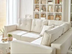 Jasny, przytulny salon z dużą, kremową kanapą. W tle białe regały pełne książek i dekoracji. Całość utrzymana w jasnej, spokojnej tonacji, gdzie dominuje **biały kolor**.