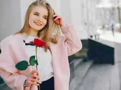 Uśmiechnięta blondynka z różą w dłoni, w różowym płaszczu. Idealna stylizacja: jak się ubrać do kina z chłopakiem, by wyglądać olśniewająco.