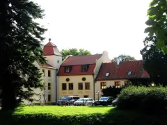 Zamek w Krokowej z czerwoną wieżą i dachami, otoczony zielenią. Przed budynkiem zaparkowane samochody.