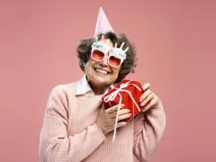 Uśmiechnięta starsza pani w okularach z napisem "Happy Birthday" i w czapce urodzinowej trzyma prezent. To dowcipne podziękowania za życzenia!