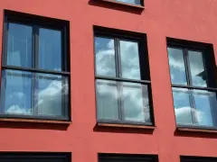 Trzy nowoczesne okna z czarnymi ramami i żaluzjami, odbijające błękitne niebo z chmurami. Wyglądają jak małe balkony francuskie.