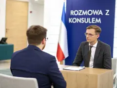 Rozmowa z konsulem. Mężczyzna w okularach i garniturze siedzi przy biurku, obok flaga.