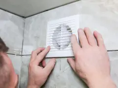 Mains d'homme ajustant une grille de VMC dans une salle de bain car une mauvaise odeur persiste.