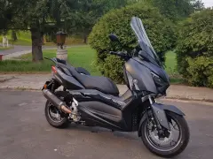 Czarny skuter Yamaha XMAX 300 zaparkowany na tle zieleni i drzew.