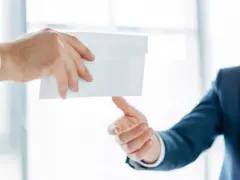 Main d'un homme en costume remettant une enveloppe blanche à une autre main. Peut-être une mise en demeure URSSAF avec un délai de paiement.