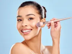Une femme souriante utilise un pinceau pour appliquer du correcteur maquillage sur sa joue.