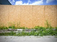 Płyta OSB na zewnątrz, otoczona dziką zielenią i fragmentami gruzu, tworzy tymczasowe ogrodzenie.