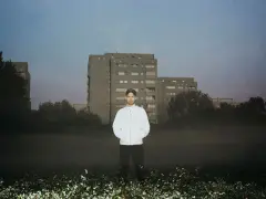 Bartosz Kruczyński stoi na tle bloków mieszkalnych, otoczony mgłą i kwitnącymi kwiatami.