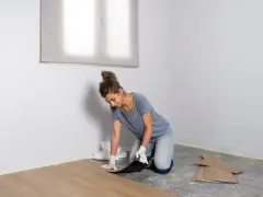Mujer con guantes instalando suelo vinílico, colocando una tabla de madera clara sobre el suelo de hormigón.