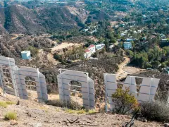 Widok zza gigantycznych liter znaku Hollywood. W oddali widać wzgórza z domami i drogami.