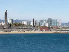 Ludzie odpoczywają na plaży, a w tle widać nowoczesne budynki Barcelony. To miasto leży nad Morzem Śródziemnym.