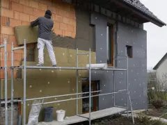 Pracownik na rusztowaniu montuje wełnę mineralną na ścianie domu. Brak cokołu na elewacji, trwa ocieplanie.
