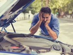 A frustrated man with hands on his head stares at his car's open hood, likely hearing a grinding noise when starting the car.