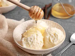 Schneeballgroße Kugeln Vanilleeis werden mit flüssigem Honig beträufelt. Ein Holzlöffel tropft goldenen Honig auf das cremige Eis.
