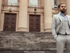 Elegancki mężczyzna w jasnej, kratkowanej marynarce i kamizelce, z chusteczką w kolorze śliwkowym w kieszeni, stoi na schodach przed budynkiem.