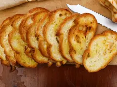 Fette dorate di pane con lievito madre, appena tostato, su un tagliere di legno. Un coltello seghettato è pronto per affettare altro pane.