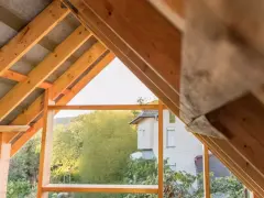 Drewniany szkielet dachu dwuspadowego w trakcie budowy. Widoczne krokwie i elementy konstrukcyjne, a w tle dom i zieleń.