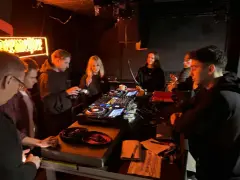 Grupa młodych ludzi przy konsoli DJ-skiej, uczących się, jak zostać DJ-em. Neonowy napis w tle.