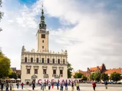 Rynek w Chełmnie z ratuszem i tablicą z napisem "Chełmno". Ludzie spacerują, podziwiając atrakcje miasta.
