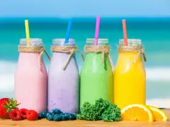 Cztery kolorowe smufi na plaży: różowe, fioletowe, zielone i żółte. Obok owoce: truskawki, maliny, borówki, pomarańcze i jarmuż.