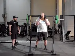 Man med skägg förbereder sig för knäböj med en safety squat bar. En tränare står bredvid.
