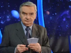 Starsza osoba w garniturze trzyma karty z napisem "1 z 10", gotowa do emisji.