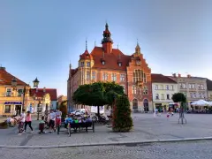 Rynek w Tarnowskich Górach tętni życiem. Ludzie spacerują, siedzą na ławkach, a dzieci bawią się na bruku. W tle widać zabytkowy ratusz.