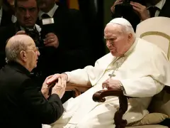 Papież Jan Paweł II w białej szacie, klęczący przed nim mężczyzna w czerni. W tle widać innych duchownych i dziennikarzy, być może z zakonu **jezuitów**.