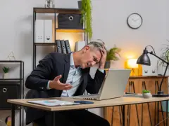 Zestresowany mężczyzna w garniturze trzyma się za głowę, patrząc na laptopa. Może właśnie stracił wadium.