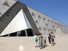 Grupa turystów podziwia nowoczesne muzeum w Gizie, którego fasada zdobiona jest hieroglifami i geometrycznymi wzorami przypominającymi piramidy.