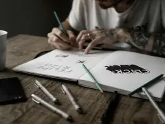 Un artiste auteur ou micro entrepreneur dessine dans son carnet, entouré de stylos, crayons et d'une tasse.