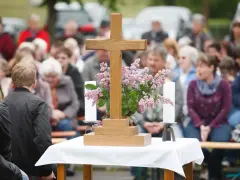 Holzkreuz mit Blumen und Kerzen vor einer Gemeinde. Ein Thema, das auch bei unterschiedlichen Konfessionen von Ehepartnern relevant ist: die Kirchensteuer.