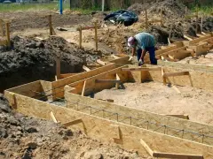 Budowa fundamentów: pracownik układa zbrojenie w drewnianym szalunku. Widać wiele desek na szalunek fundamentu, gotowych do zalania betonem.