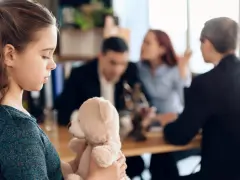 Smutna dziewczynka z misiem w ramionach, w tle rodzice i kurator procesowy dyskutują.
