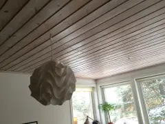 En vit lampskärm hänger från ett tak av träpanel. En tunn sladd dras i taket för att ansluta lampan.