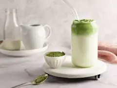 Pyszny matcha latte w szklance, obok miseczka z zielonym proszkiem i łyżeczka.