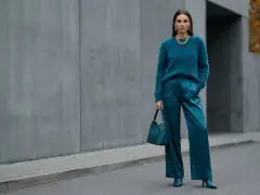 Kobieta w eleganckim, futrzanym swetrze i satynowych spodniach w kolorze petrol. Torebka i buty również w tym samym odcieniu.