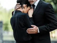 Smutna kobieta w czarnej sukience przytula się do mężczyzny w garniturze. On obejmuje ją ramieniem, pocieszając w trudnym momencie. Pytanie, jaki krawat na pogrzeb, jest tu nieistotne.