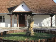 Bronowicka chata symbol: snop zboża przed tradycyjnym domem z drewnianymi elementami i ceramicznym dachem.