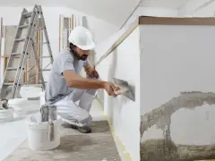 Remont w bloku w niedzielę? Mężczyzna w kasku tynkuje ścianę, obok drabina i wiadra z materiałami.