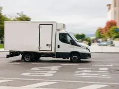 Biała chłodnia, kategoria C1, pędzi ulicą. W tle zamazane drzewa i budynki.