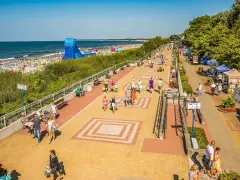 Tętniąca życiem ustka promenada w słoneczny dzień. Ludzie spacerują, odpoczywają na ławkach, a w tle widać plażę pełną turystów i morze.