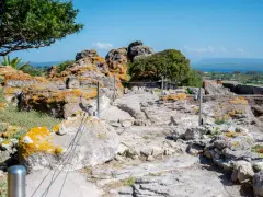 Ancient Italian ruins with weathered stones covered in orange lichen, a pathway winding through the site, and a distant blue sea under a clear sky.