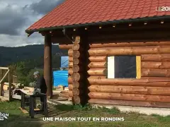Une maison en rondins de bois en construction, avec un homme travaillant à proximité. Le toit est rouge et le paysage est montagneux.