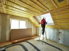Operaio in piedi su una scala per isolare il tetto di una mansarda con materiale isolante giallo.