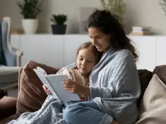Mama czyta dziecku bajkę, otuleni kocem. Widać, ile jest opieki na dziecko w tej chwili.